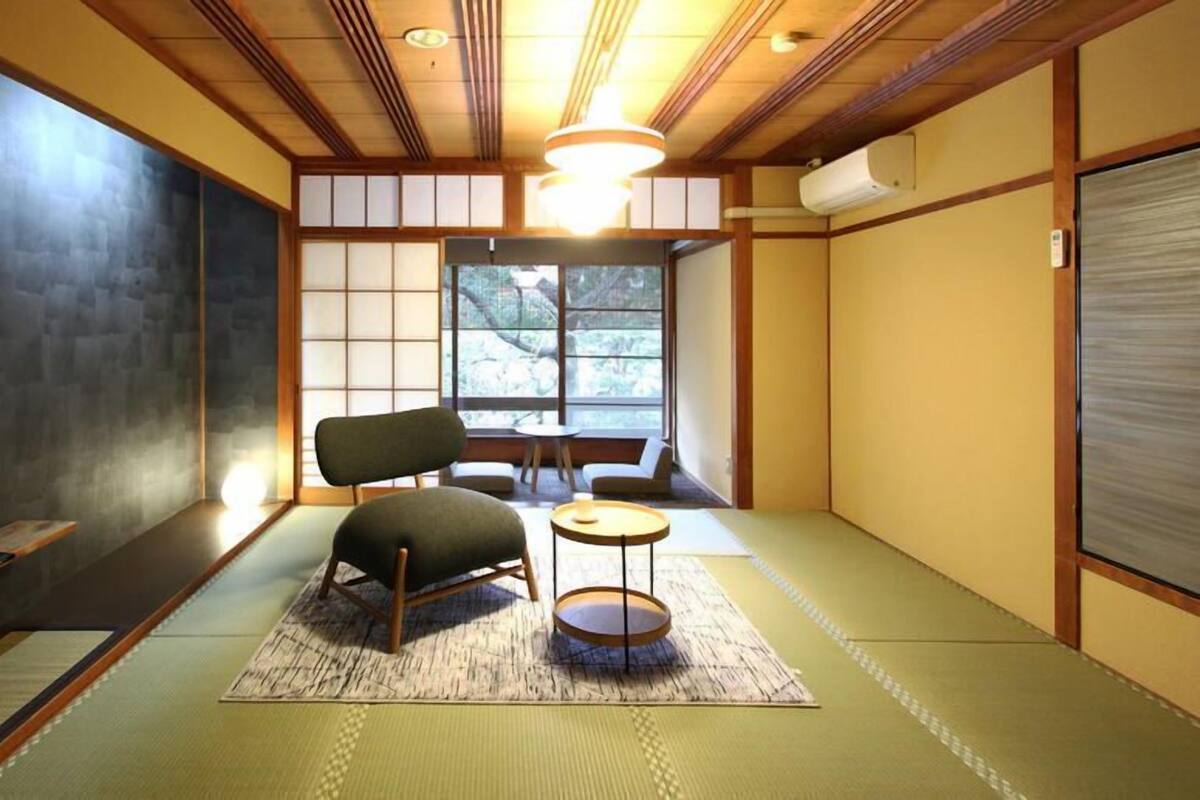 Une pièce traditionnelle avec un sol recouvert de tatamis, une petite table, une chaise et une fenêtre donnant sur des arbres.