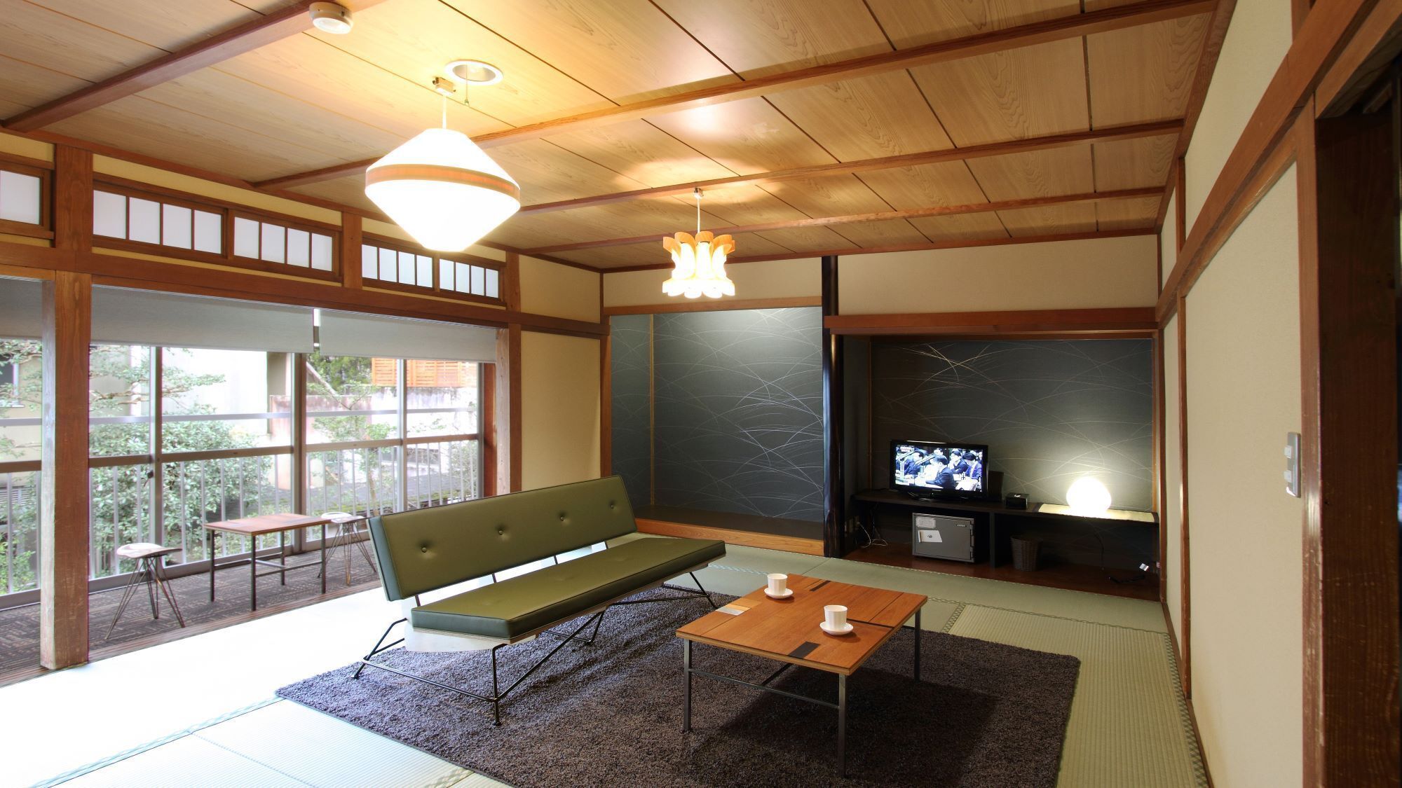 Una camera in stile tradizionale giapponese, con pavimento in tatami, un divano verde basso, un tavolino in legno e un televisore su una parete scura.