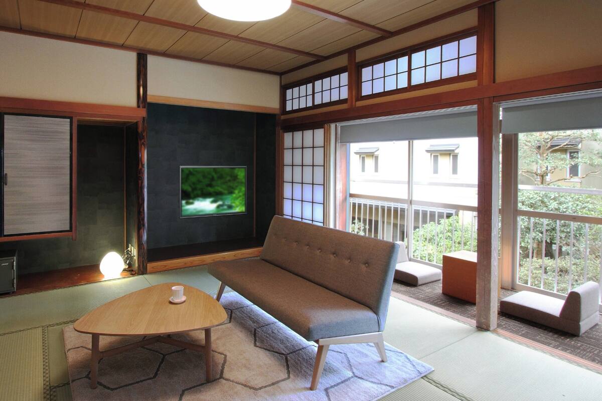 Une pièce de style japonais traditionnel, avec un sol recouvert de tatamis, une table basse, un canapé, un téléviseur, des portes coulissantes et une vue sur l’extérieur.