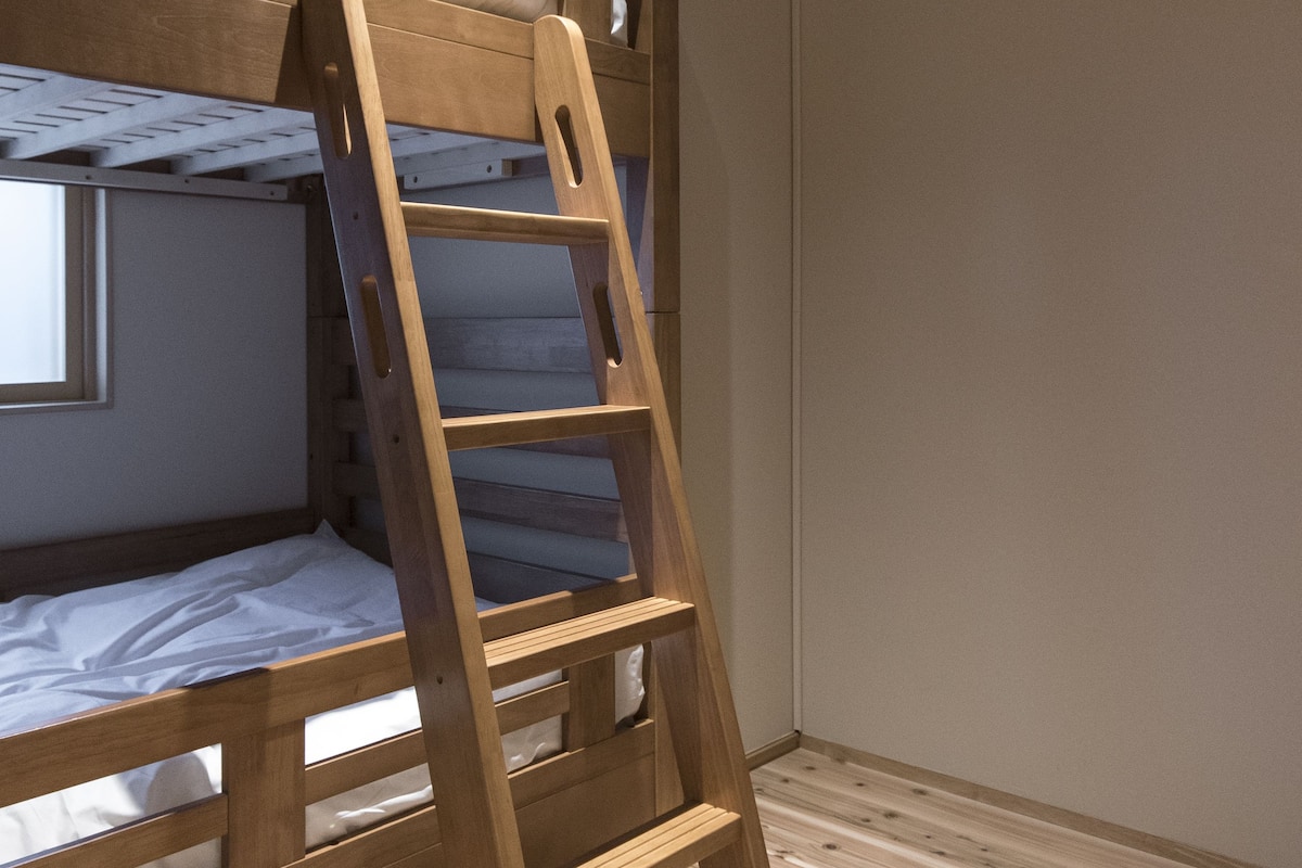 Een stapelbed met een houten trap, een raam en een airconditioning op de muur.