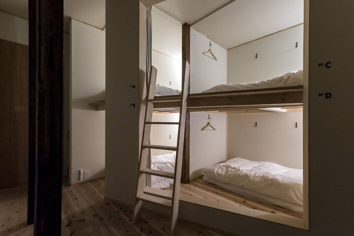 Een studentenkamer met stapelbedden, een houten trapje en een witte muur met aanduidingen.