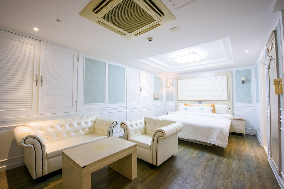 Una habitación de hotel con una cama, dos sofás blancos, una mesita y una ventana con persianas.
