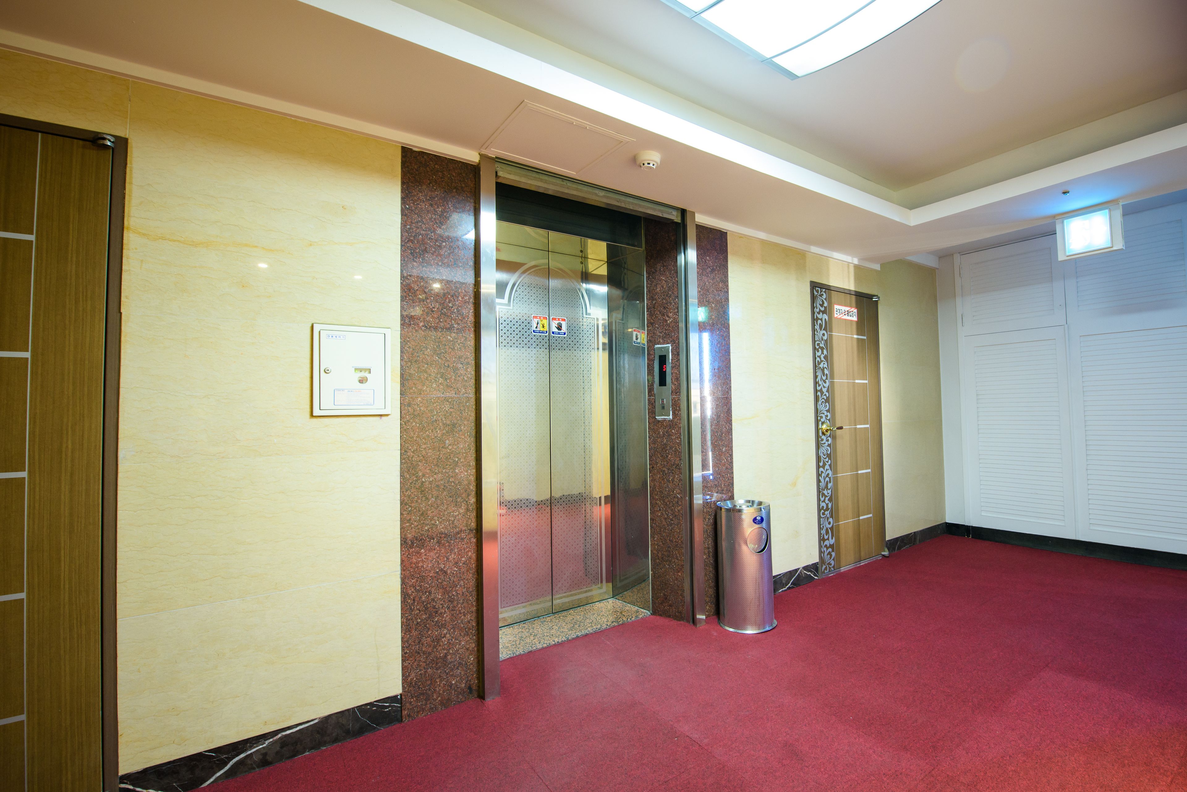 An elevator in a hallway with a red carpet, a trash bin, and a security camera.