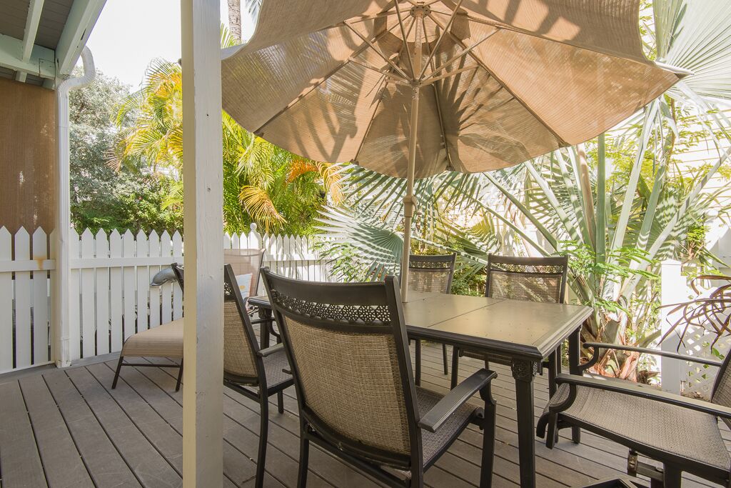 A covered outdoor patio with a table and chairs, surrounded by lush tropical plants and a white picket fence.