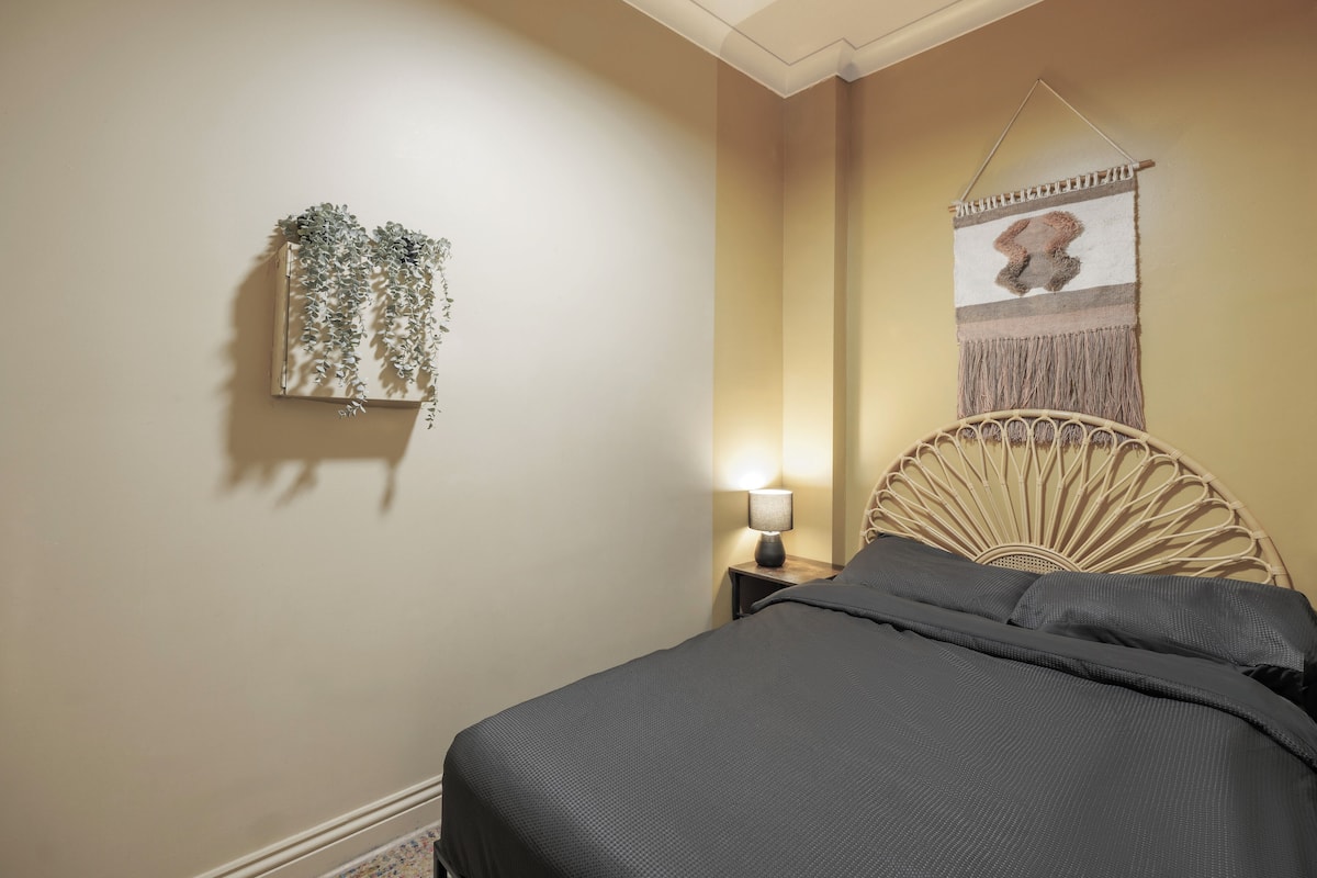 A neatly made bed with a dark gray bedspread, a wall-mounted plant, a wall hanging, and a bedside lamp.