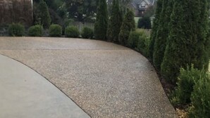 Un chemin en béton incurvé, doté d’une surface texturée, bordé d’arbustes et d’arbres aux formes soigneusement taillées.