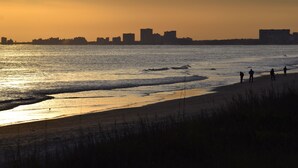 Ein Strand mit Menschen, die am Ufer entlanggehen, einer Stadtlandschaft im Hintergrund und der untergehenden Sonne über dem Wasser.