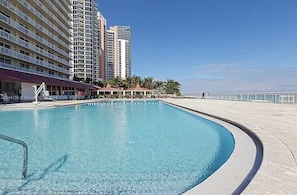 A swimming pool with a curved edge, surrounded by a concrete deck. In the background, there are high-rise buildings and a clear sky. - Beautiful Hotel Studio on the beach. STR-01535 (Sunny Isles Beach)