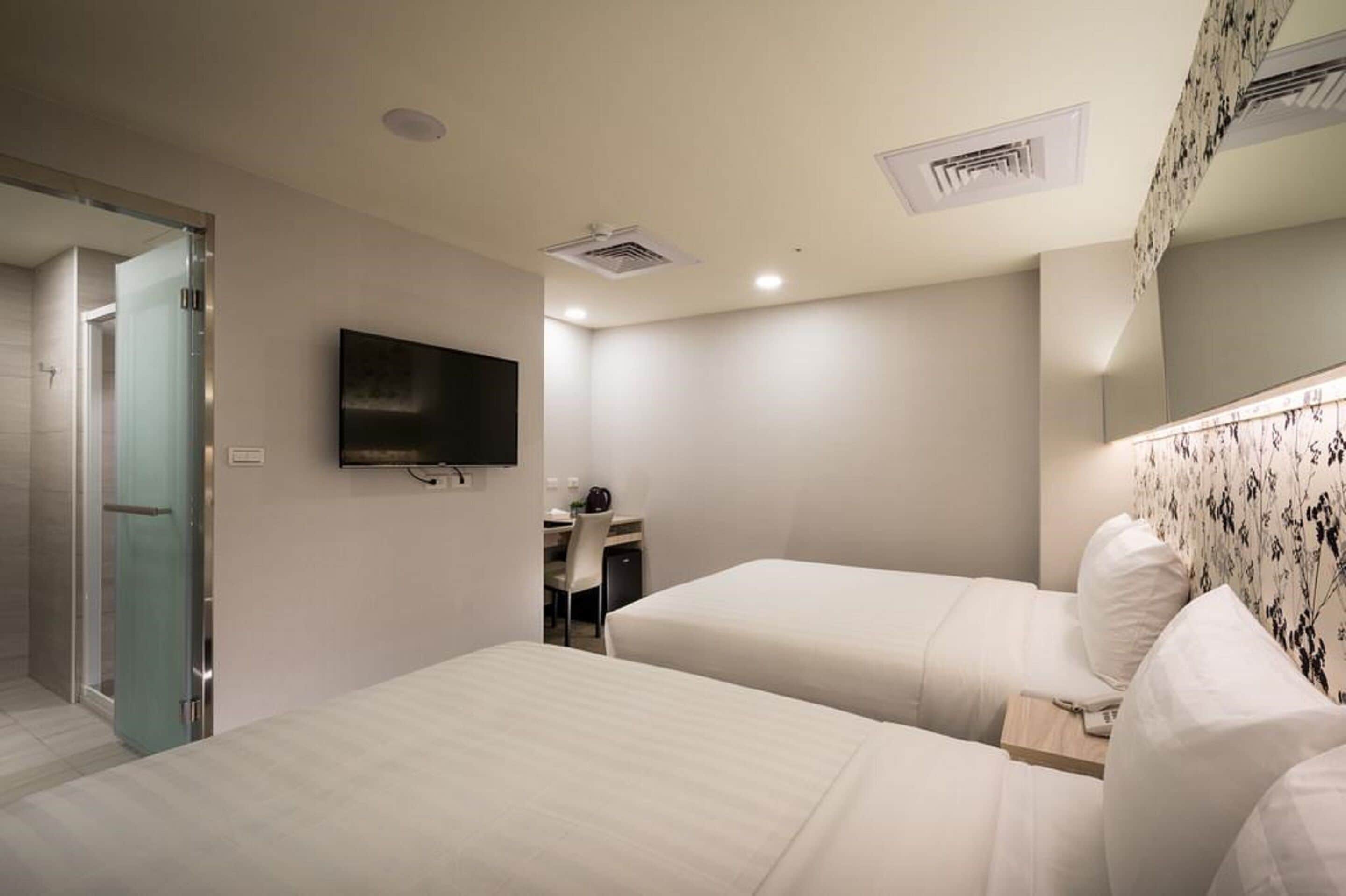 A hotel room with two beds, a desk with a chair, a flat-screen TV mounted on the wall, and a shower area.