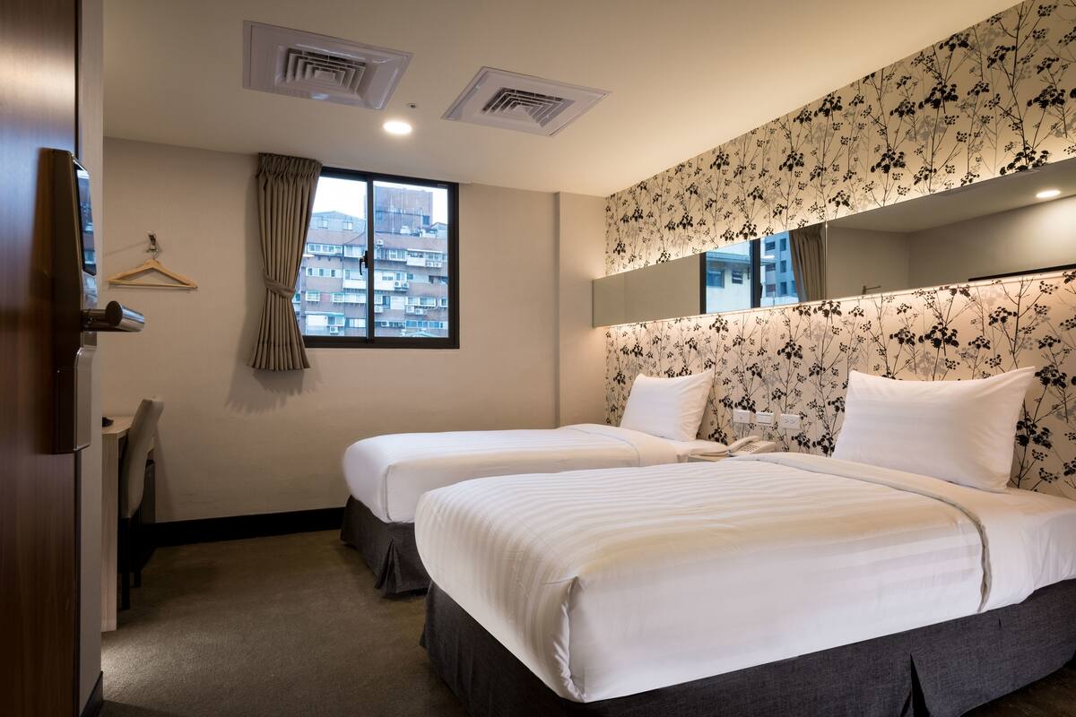 A hotel room with two beds, a window with a view of buildings, a hanging coat rack, and a floral-patterned headboard.