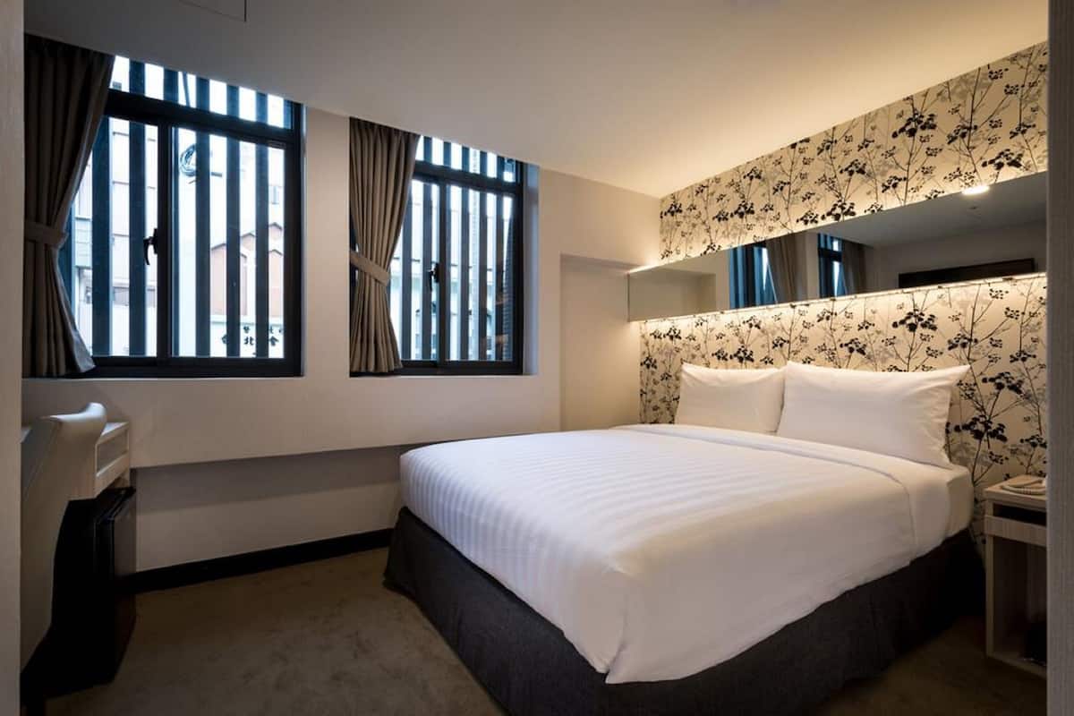 A hotel room with a large bed, two windows with curtains, a bedside table, and a wall-mounted mirror.