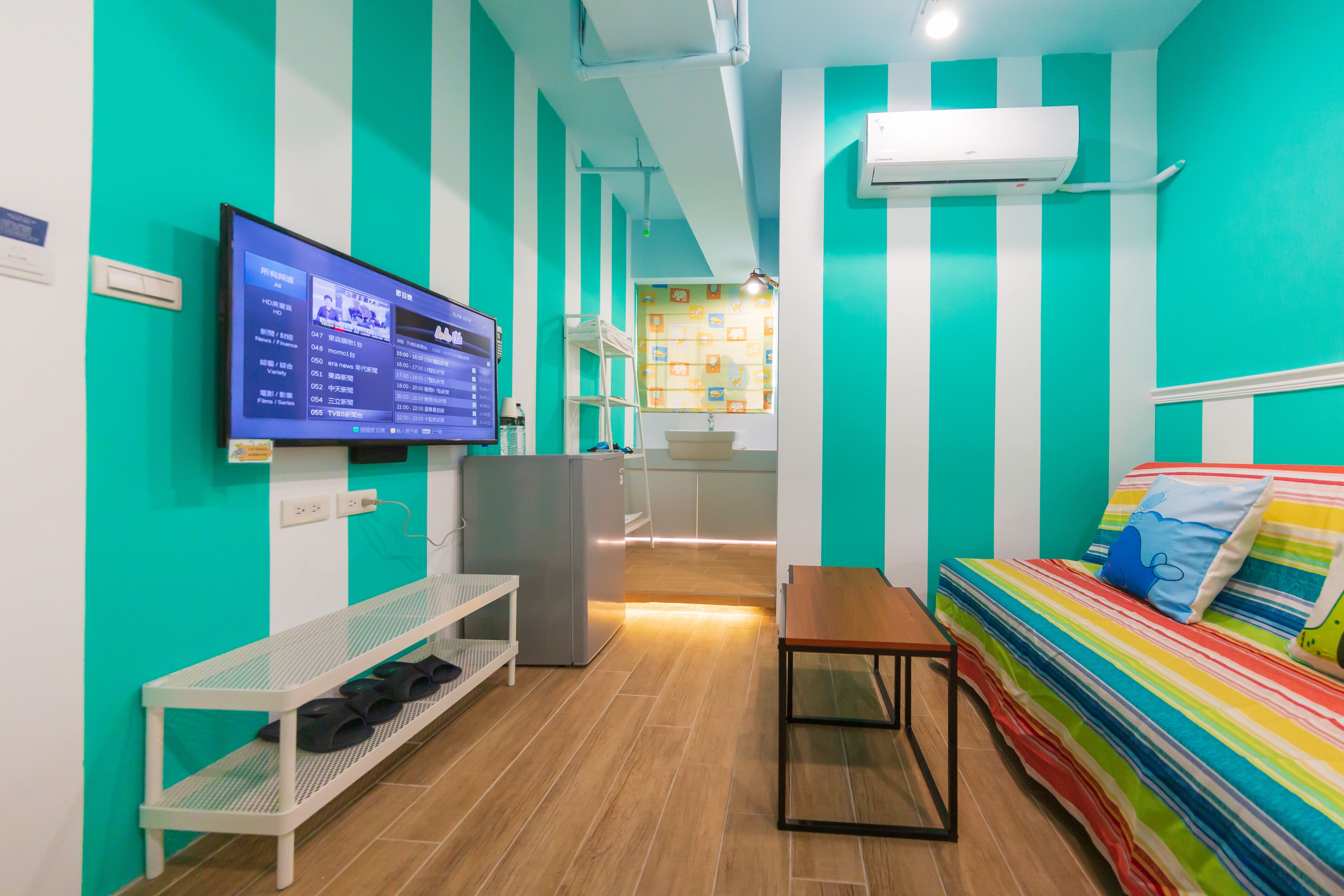 A room with teal walls, a bed with colorful bedding, a TV on the wall, a small table, and a bench with shoes.