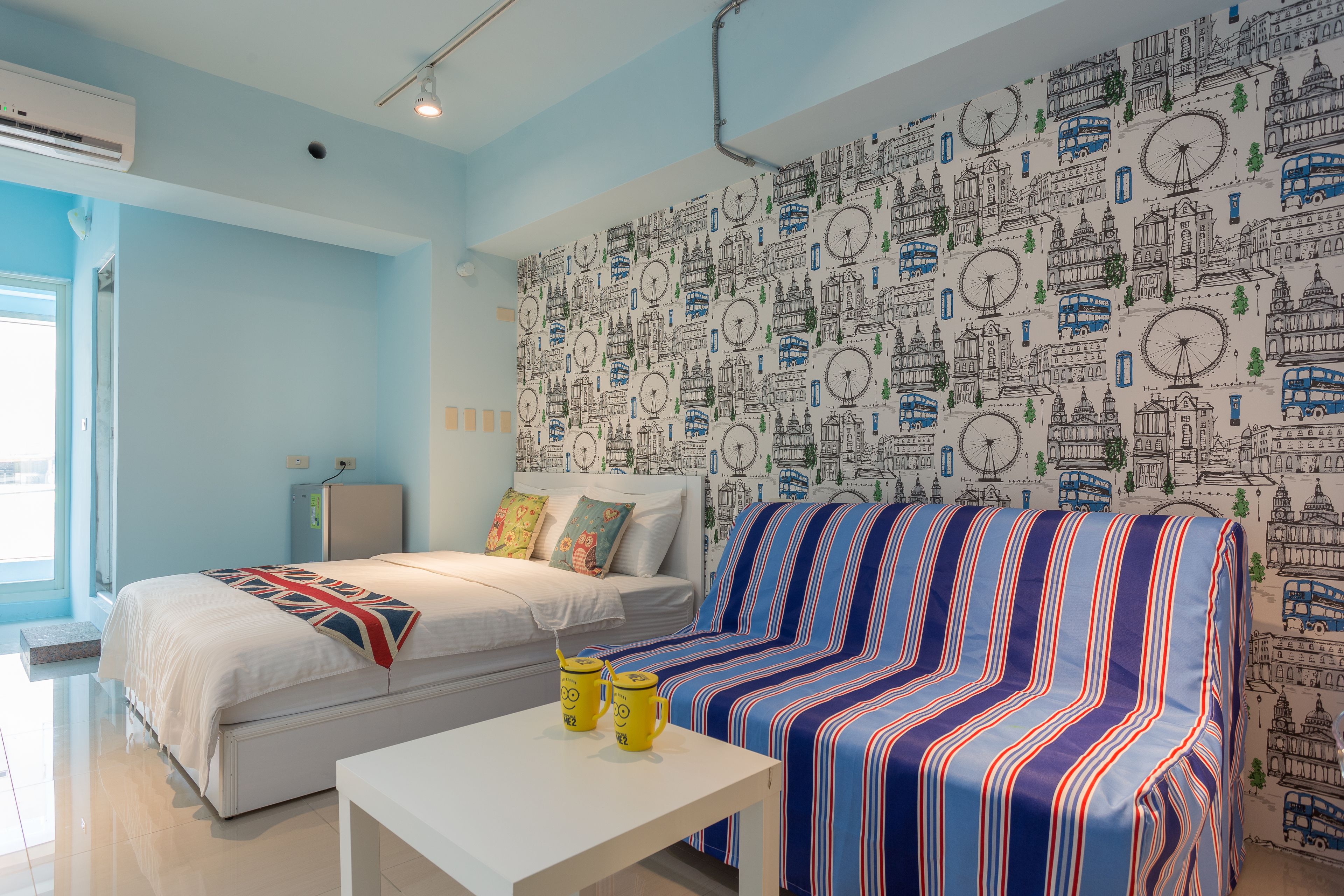 A room with a bed, a sofa, a small table, and a wall with a cityscape pattern.