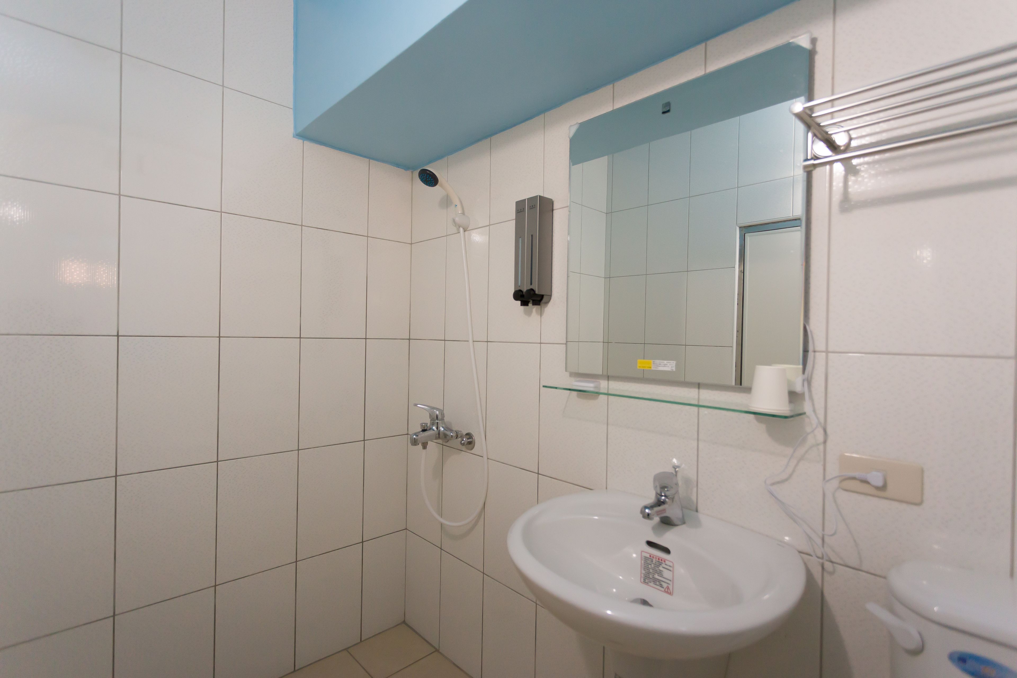 Une salle de bain avec des carreaux blancs, une étagère en verre au-dessus du lavabo, une douchette à main et des toilettes.