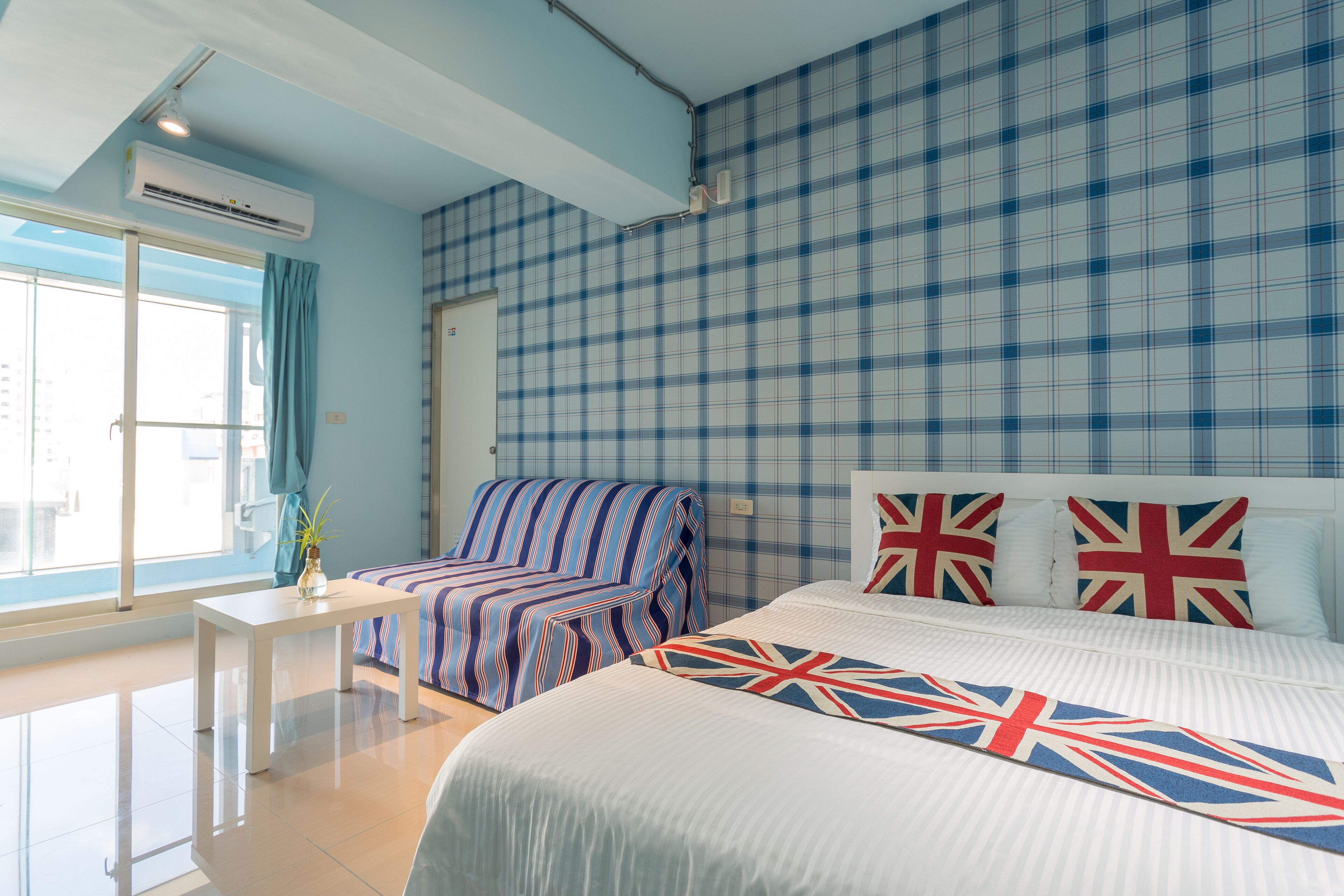 A hotel room with a checkered blue and white wall, a bed with a Union Jack pillow, a striped armchair, a small white table with a vase, and a window with curtains.
