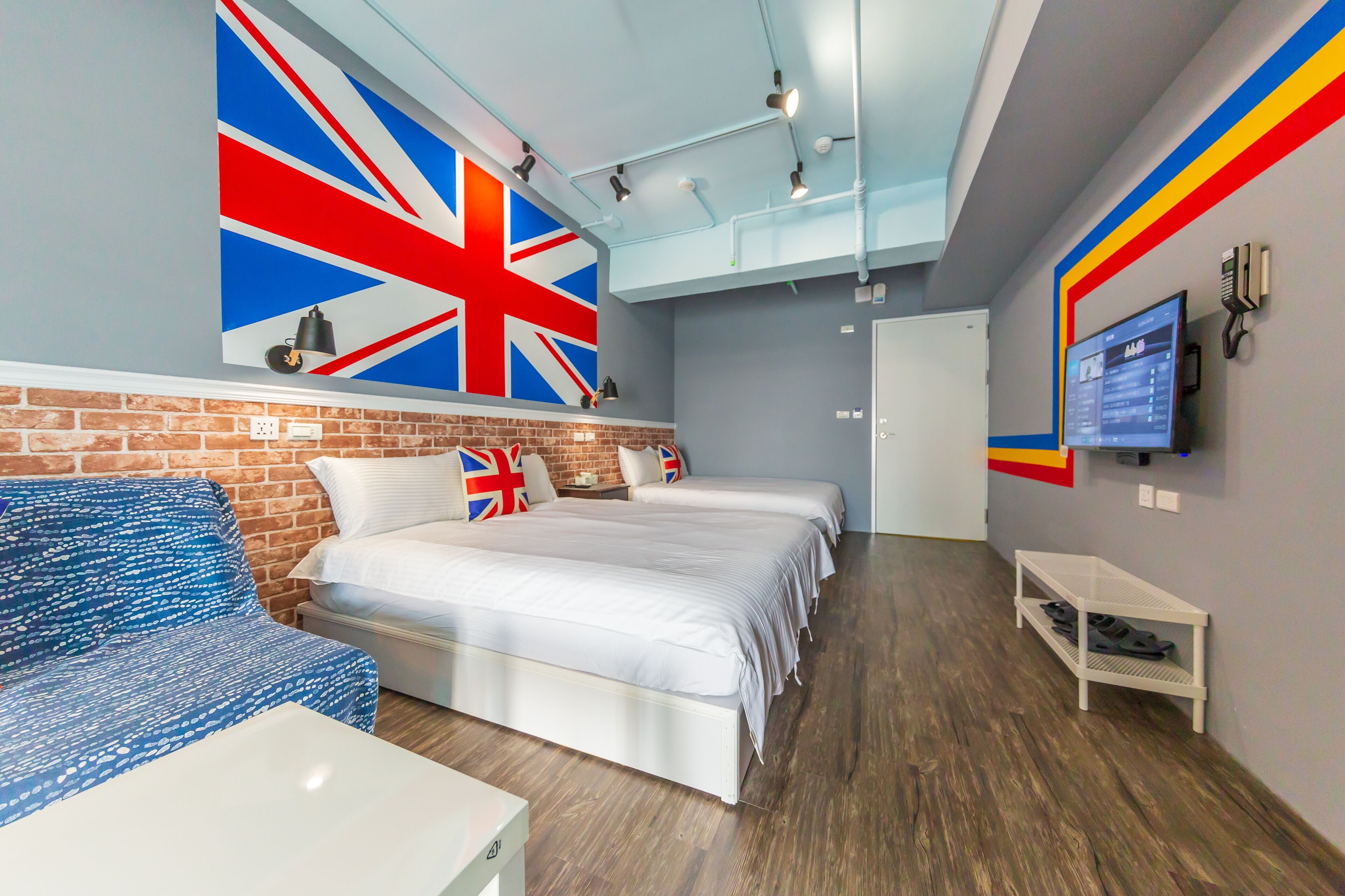 A hotel room with a brick wall, two beds with white bedding, a blue patterned sofa, a TV on the wall, and a shelf with shoes.