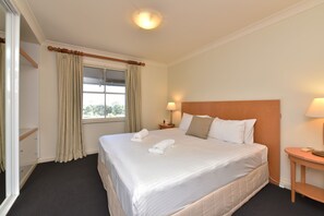 Habitación de hotel con una cama grande, una mesita de noche con lámpara, una ventana con cortinas y una puerta.