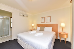 A hotel room with a large bed, two bedside tables with lamps, a wooden headboard, an air conditioning unit, and a glass shower enclosure.