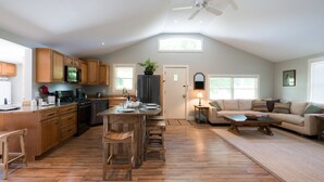 A spacious living area with a kitchen, wooden flooring, a ceiling fan, and a large window providing natural light. - The Red Hen Cottage-Asheville-with Hot Tub and EV charger! (Asheville)