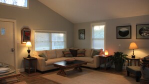 A living room with a sofa, coffee table, and lamps. There are windows with blinds, a door, and a wall with framed pictures. - The Red Hen Cottage-Asheville-with Hot Tub and EV charger! (Asheville)