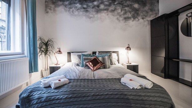 Ein modernes Schlafzimmer mit einem großen Bett, zwei Nachttischen, einem an der Wand montierten Fernseher und Blick auf die Stadt durch das Fenster.