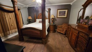 Ein Bett mit Baldachin, ein Holzkopfteil, eine Holzkommode mit Schubladen, ein kleiner Holzstuhl und ein gerahmtes Bild an der Wand.