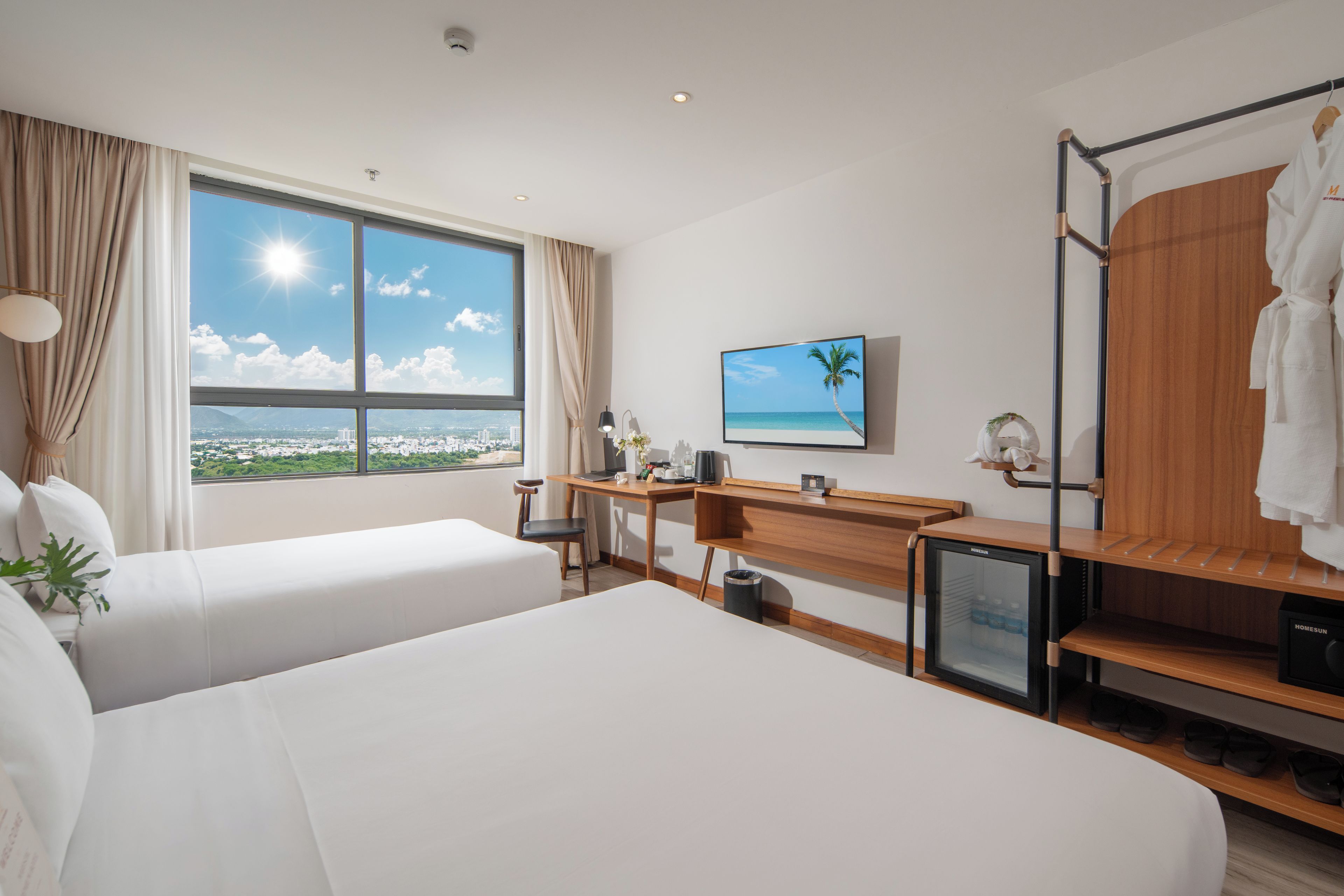 A hotel room with two beds, a desk, a small fridge, a TV, and a view of the city and sea.