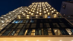 Entrada de hotel con diseño moderno, que incluye una escalera hacia el lobby, un mostrador de recepción y un cartel de bienvenida para los huéspedes.