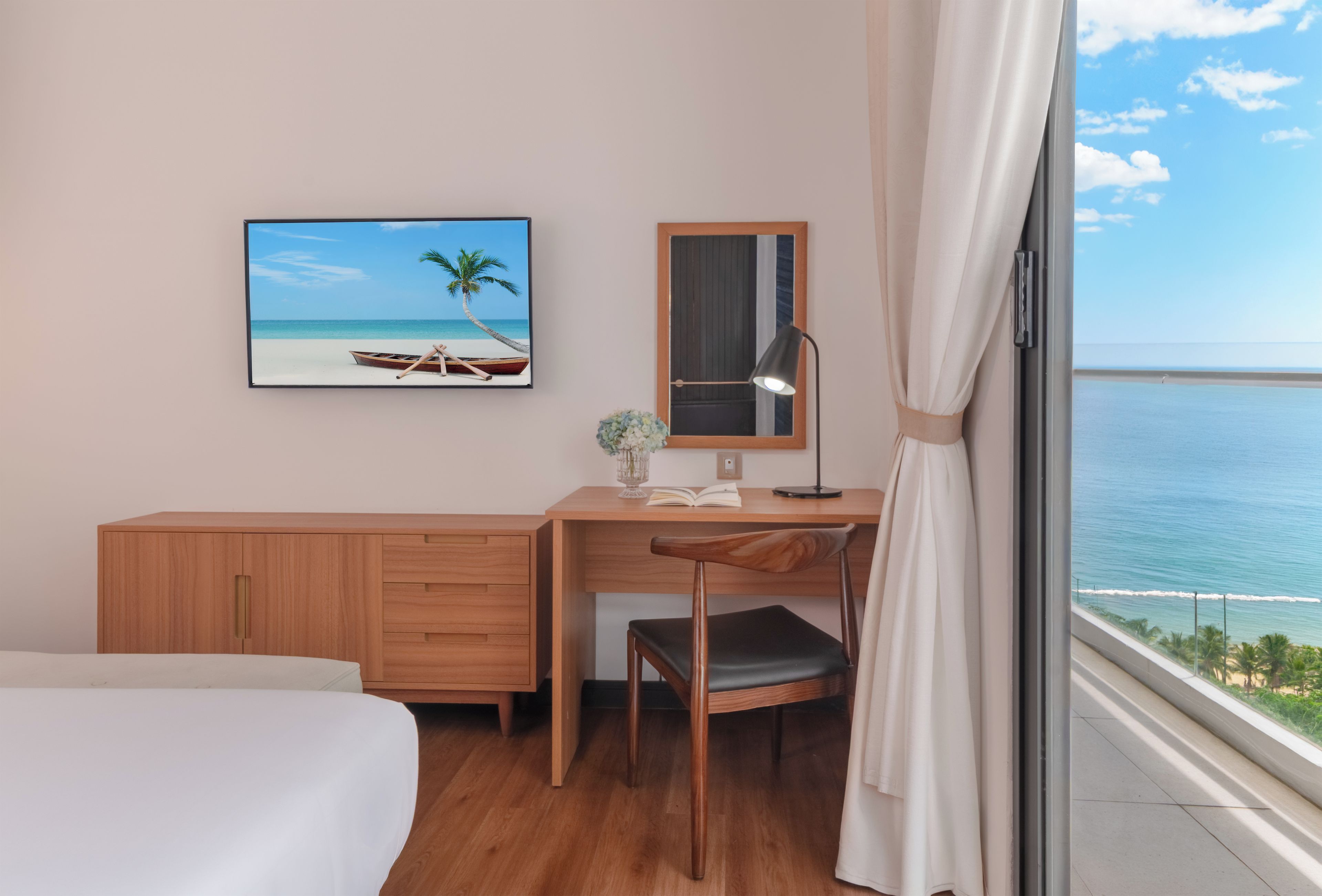 A hotel room with a bed, a desk, a chair, a desk lamp, a mirror, a framed picture of a beach, and a view of the ocean.