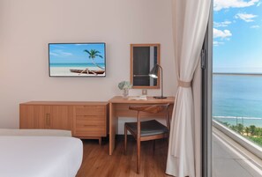A hotel room with a bed, a desk, a chair, a desk lamp, a mirror, a framed picture of a beach, and a view of the ocean. - Majestic Premium Hotel (Nha Trang)
