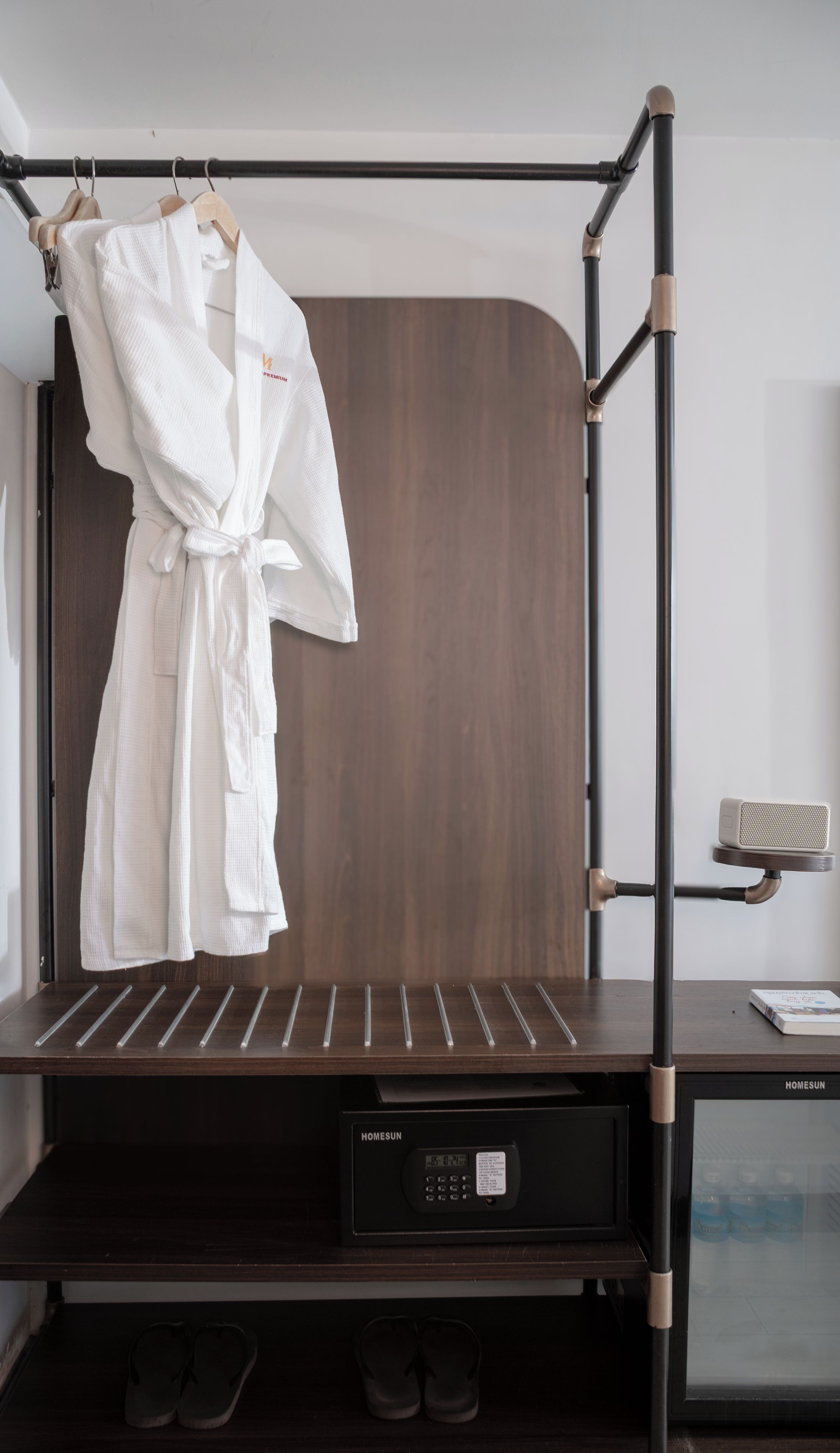 Habitación de hotel con un armario de madera oscura, estructura de cama metálica negra, bata blanca colgada en el armario, microondas y una pequeña mesa con un libro.