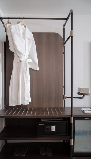 Habitación de hotel con un armario de madera oscura, estructura de cama metálica negra, bata blanca colgada en el armario, microondas y una pequeña mesa con un libro.