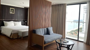 Una habitación de hotel moderna con cama, sofá, una mesita de cristal y un amplio ventanal con vistas a la playa y al océano.
