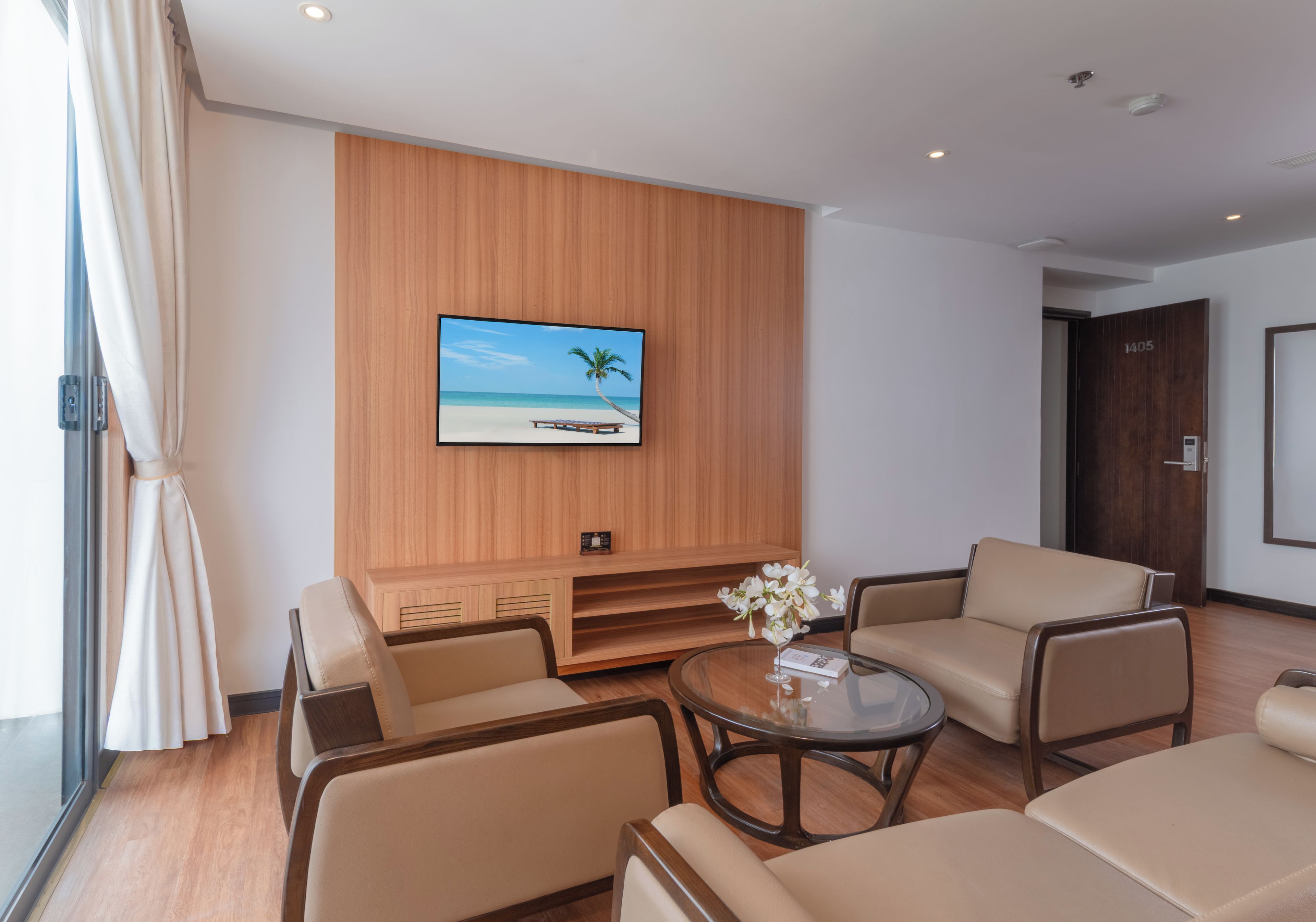 Una sala de estar moderna con un televisor de pantalla plana grande montado en una pared de paneles de madera, una mesa de centro de vidrio con un jarrón de flores blancas y dos sofás color beige.