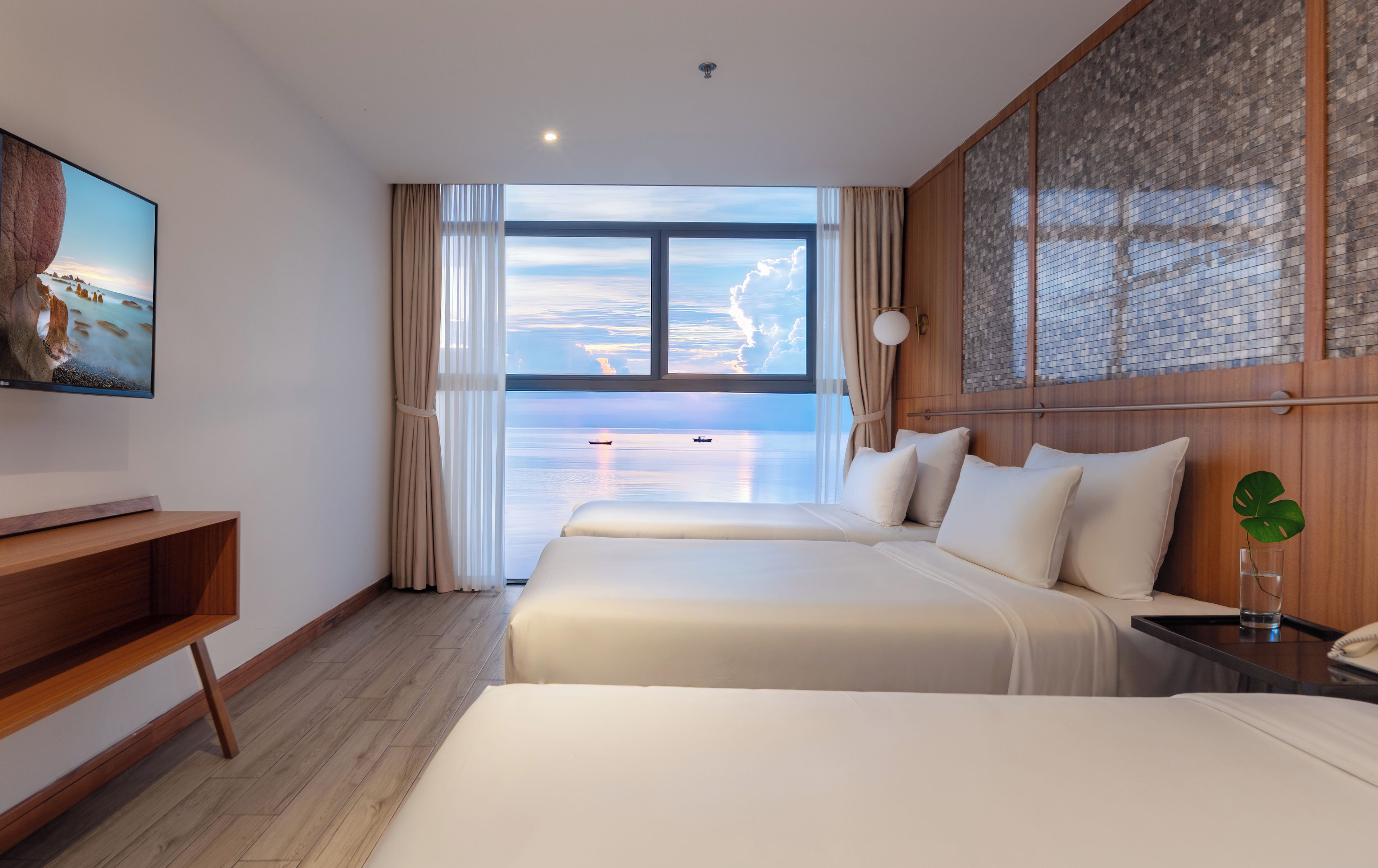 A hotel room with a large window, two beds, a TV, and a wooden desk.