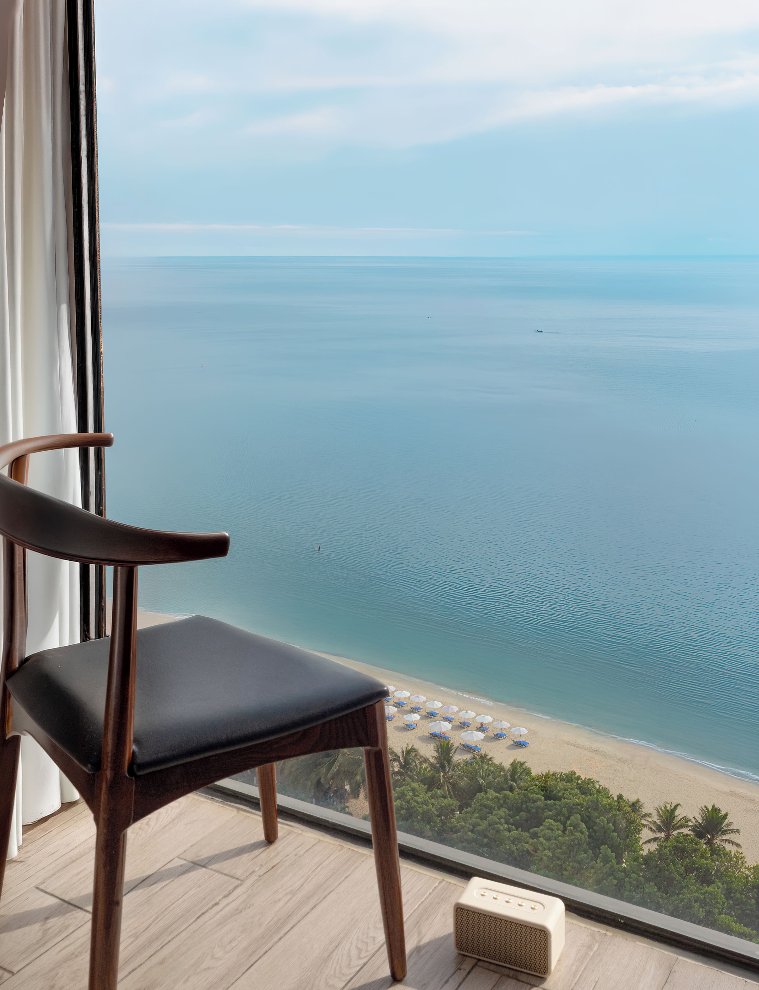 Una silla de madera junto a una ventana con vistas a una playa con sombrillas y palmeras.