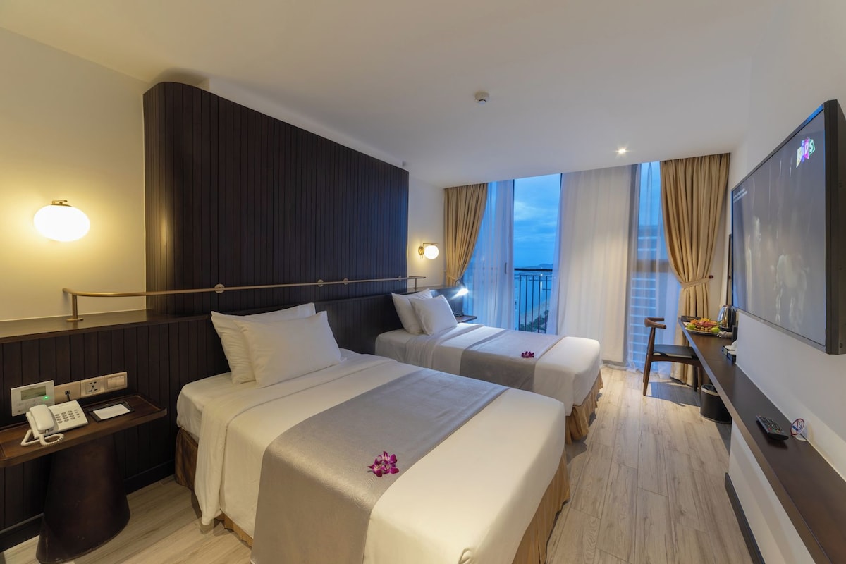 Une chambre d’hôtel avec deux lits, un bureau avec un téléphone, une télévision et un balcon avec des rideaux.