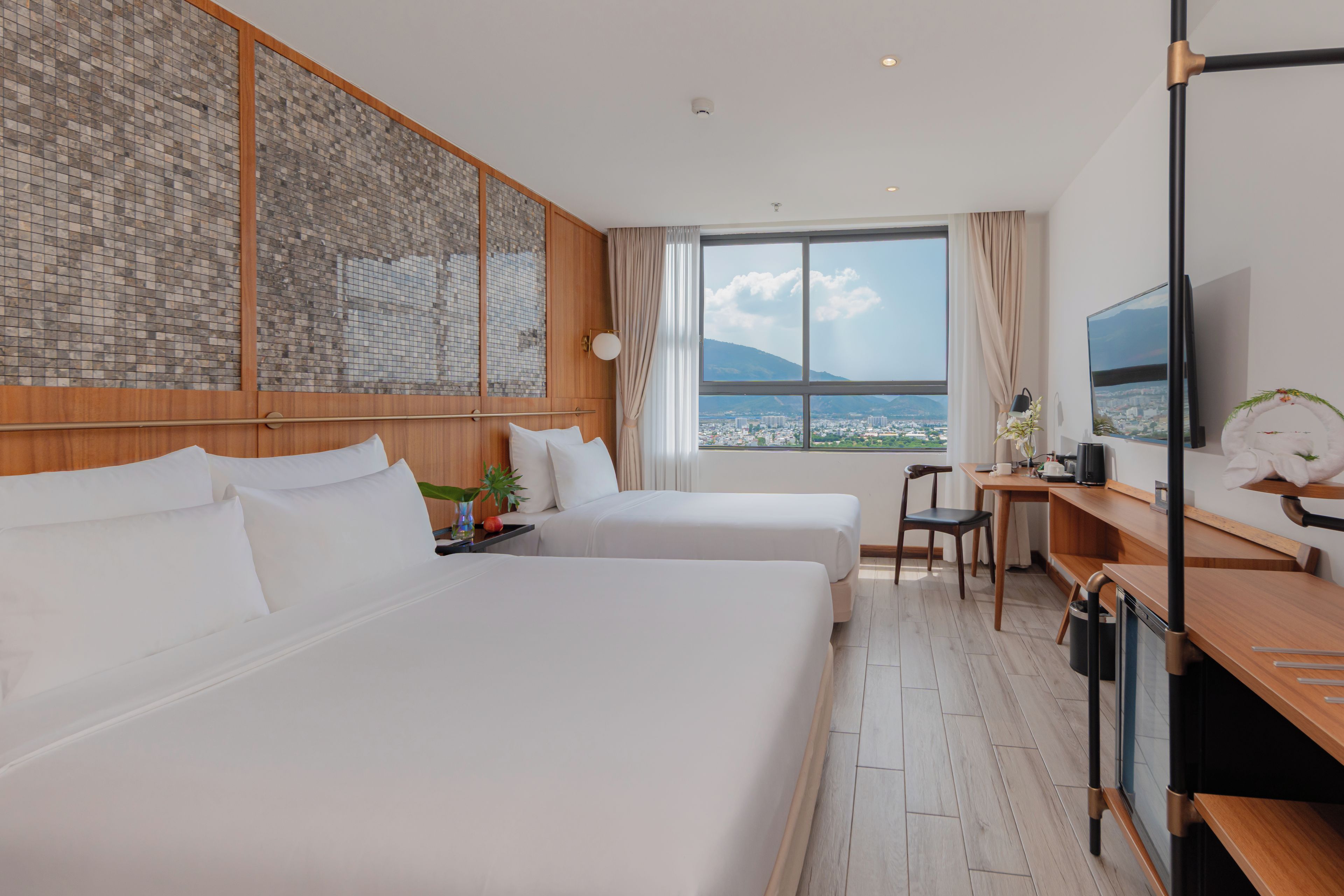 Habitación de hotel con dos camas, un escritorio, un televisor y un amplio ventanal con vistas a la ciudad.
