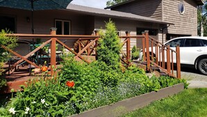 Une maison en bois avec un patio couvert, une voiture garée devant et un jardin agrémenté de diverses plantes et fleurs.