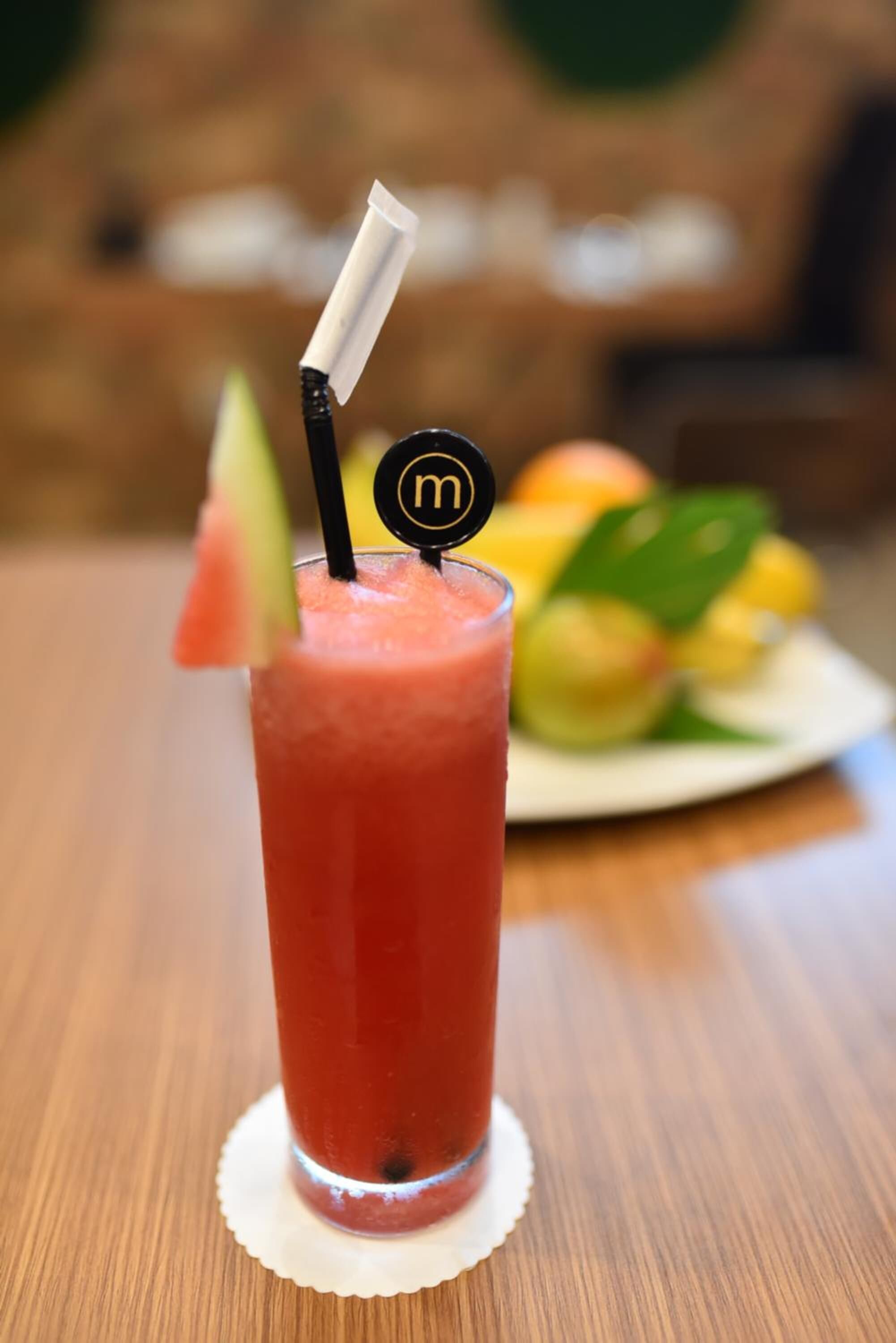A tall glass of red beverage with a straw and a slice of watermelon, placed on a wooden table.