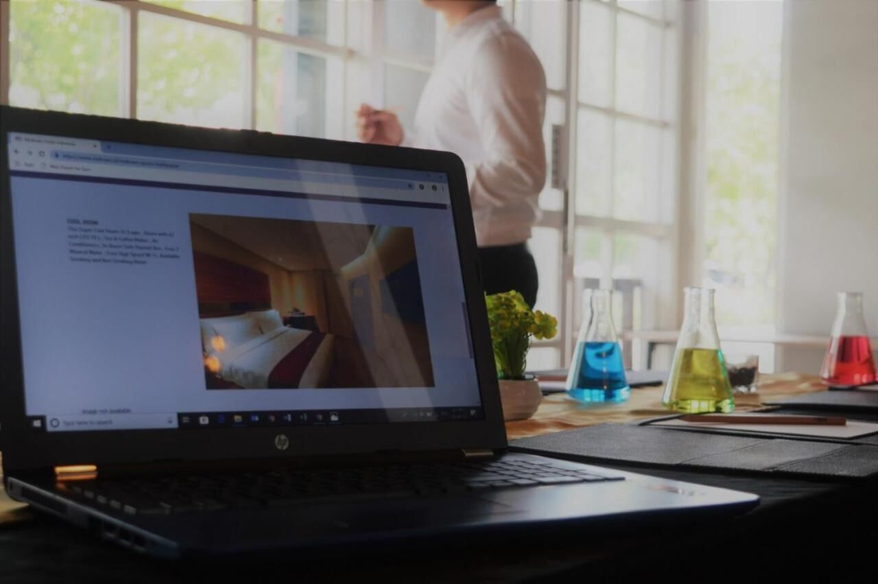 Una computadora portátil sobre un escritorio con vista al exterior, una persona al fondo y cristalería de laboratorio en la mesa.