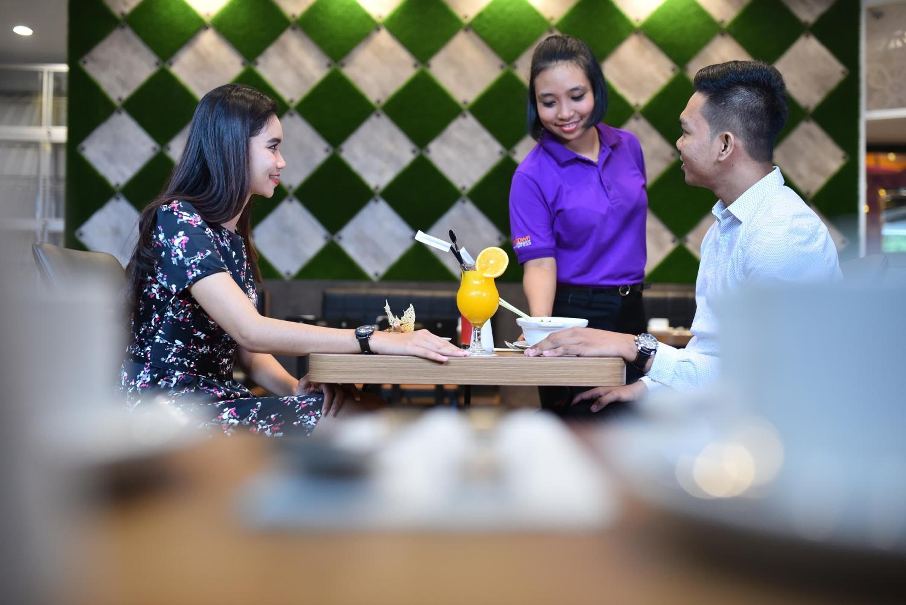 A restaurant setting with a table, a drink with a lemon slice, and a person in a purple shirt serving.