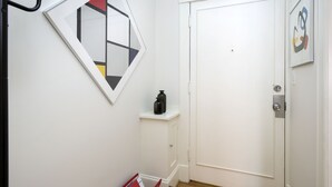 Un pasillo con una puerta blanca, un mueble blanco pequeño con dos botellas negras, y una obra de arte abstracta enmarcada en la pared.