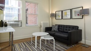 Una sala de estar moderna con un sofá de cuero negro, una mesa de centro blanca, una mesita auxiliar pequeña y un tapete a rayas. Hay cuadros enmarcados en la pared y una lámpara de pie.