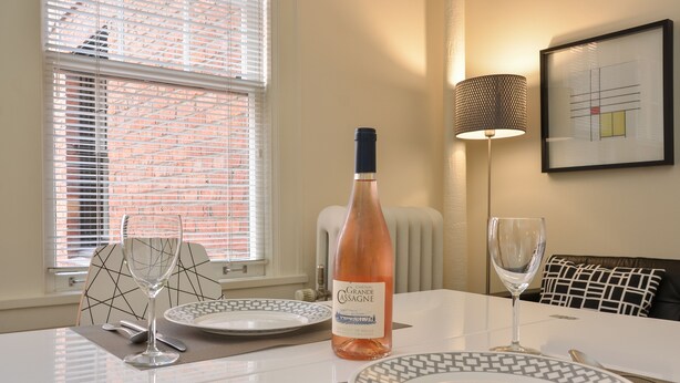 Una mesa de comedor preparada con platos, cubiertos y una botella de vino, con una ventana y persianas al fondo.