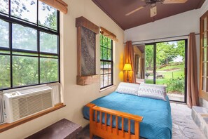 A bedroom with a large window, a bed with blue bedding, a wooden headboard, and a view of a garden. - The Guadalupe River right in your backyard! River living at its best! (New Braunfels)