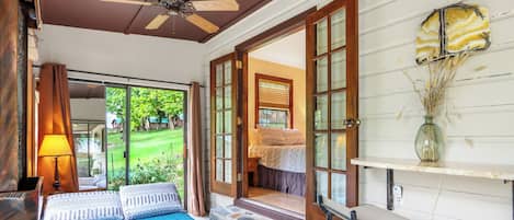 A bedroom with a bed, a desk, and a chair. There is a window with curtains, a ceiling fan, and a vase with dried plants on a table.