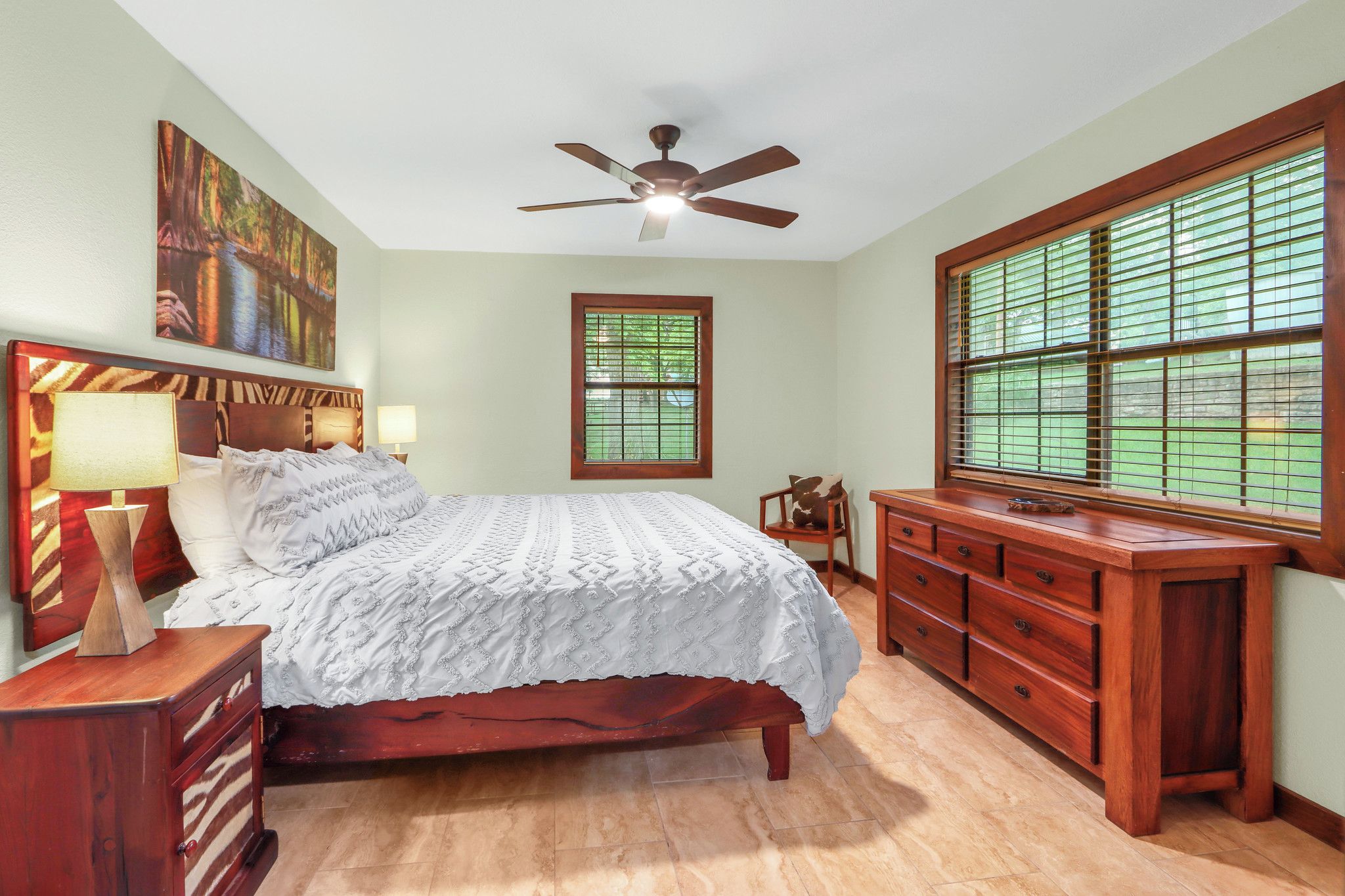 A bedroom with a wooden bed, a nightstand, a dresser, a ceiling fan, a window with blinds, and a painting on the wall.