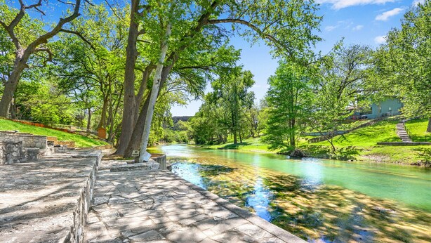 Un sendero de piedra que conduce a un río, con un pequeño puente y una zona cubierta de césped y árboles.