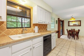 A kitchen with a sink, dishwasher, white cabinets, a window, and a dining area with a table and chairs.